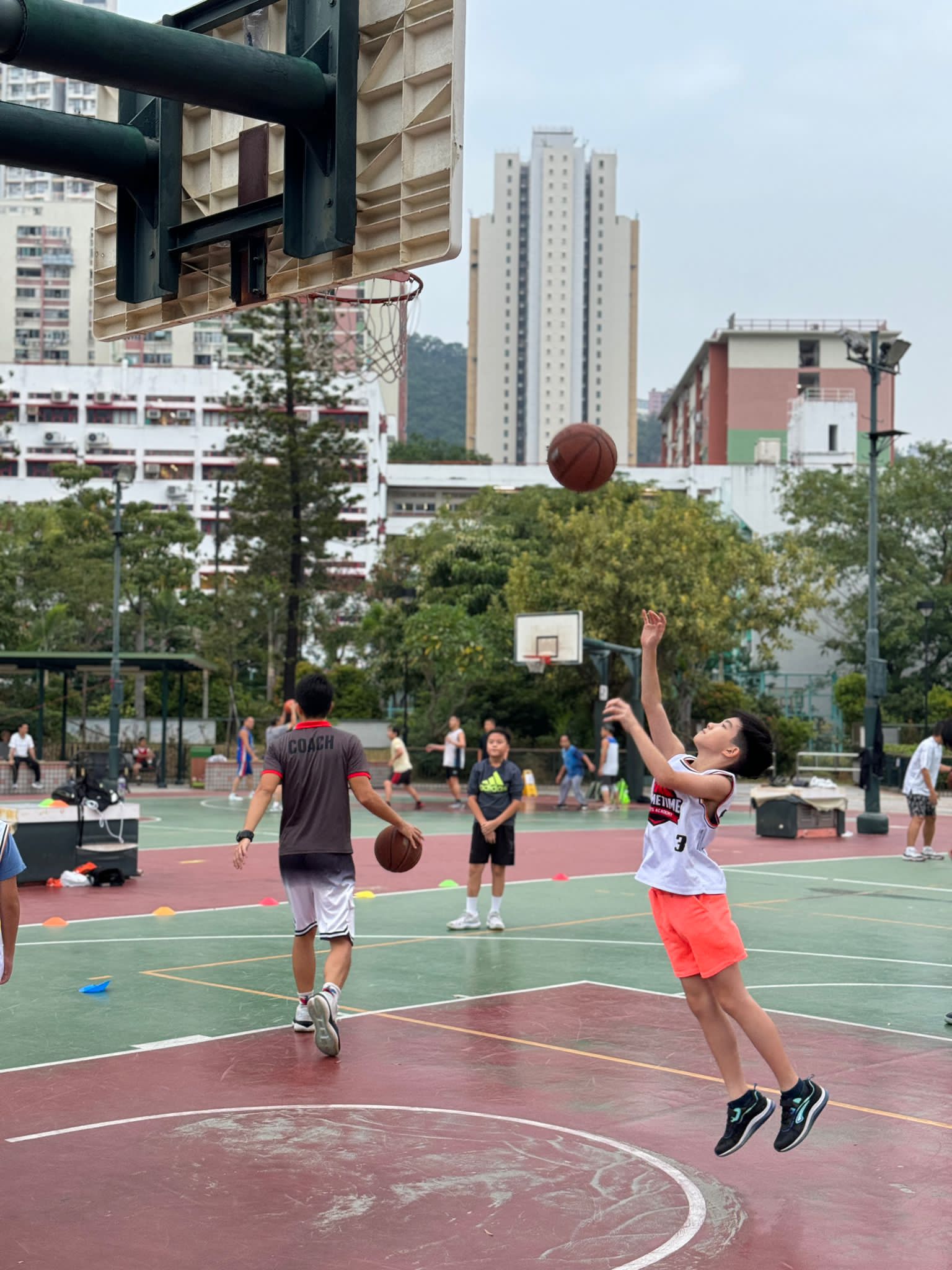 A person wearing an "11" jersey jumps to shoot a basketball on an outdoor court while others practice and a coach observes, all forming part of an intense personal development training program.