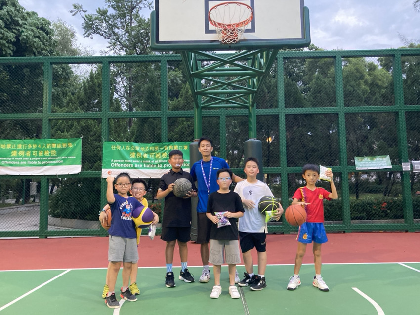A group of kids and an adult stand on a basketball court holding basketballs and snacks. A basketball hoop is in the background. Trees and green fencing are seen behind them, creating the perfect setting for personal growth through basketball training.
