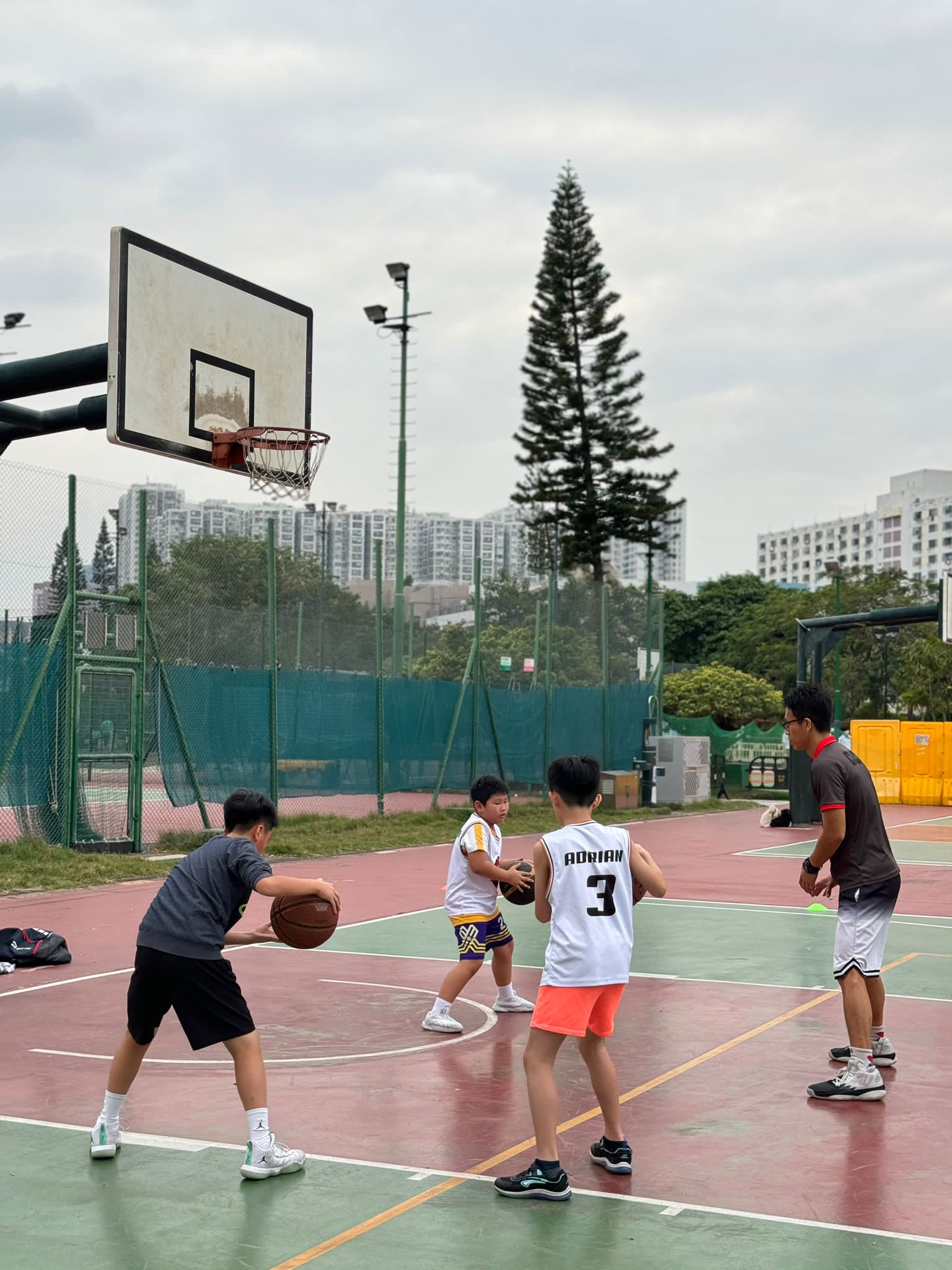 A group of four individuals, including children and an adult, play basketball on an outdoor court. One child is dribbling the ball while the others watch, and the adult appears to be coaching as part of a structured basketball training program.