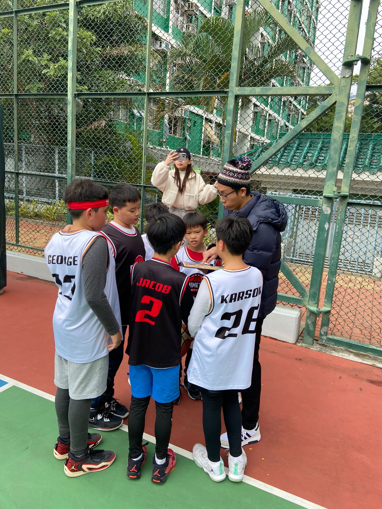 A group of young boys in basketball jerseys huddle with an adult on an outdoor basketball court in Hong Kong, while another person captures the moment, illustrating their journey of personal growth through dedicated basketball training.
