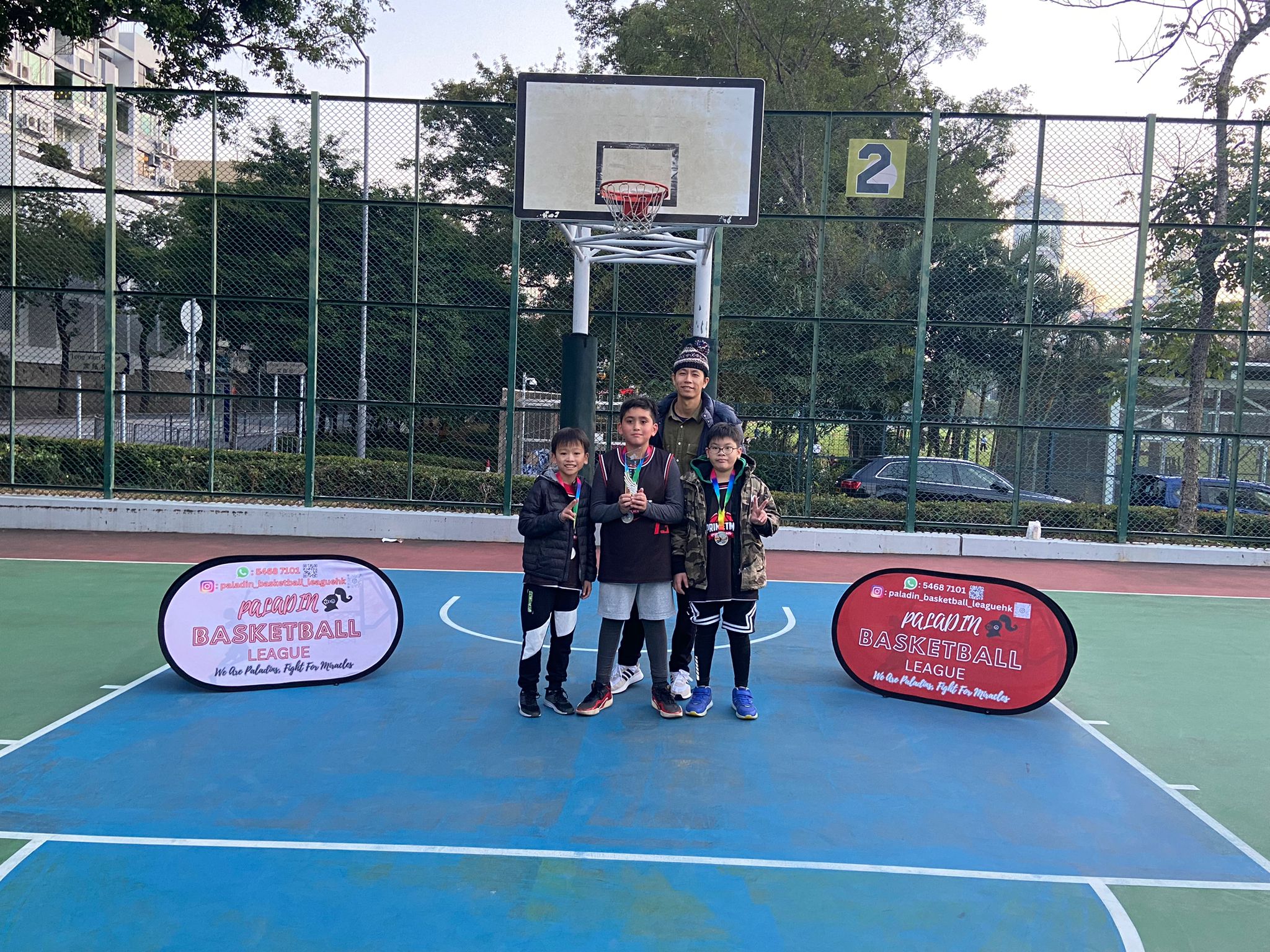 Four children and an adult standing on an outdoor basketball court, holding medals. Two signs reading "Kowloon Basketball League" are on either side of the group. A basketball hoop is visible in the background.